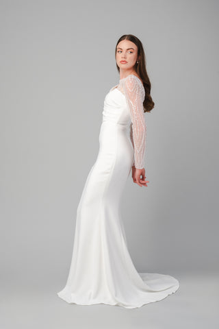 The Elea Dresses Alexia dress in white features a diagonally pleated top with long sheer sleeves embellished with stones. Designed to hug the body, this dress creates a flattering silhouette. A small train at the back adds a refined and dramatic touch.