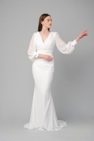 The Elea dresses Clara dress in white features a pleated V-neckline and is tailored around the waist. It is a fitted dress with a small train. The sleeves are balloon-styled, and the cuffs feature detailed buttons.