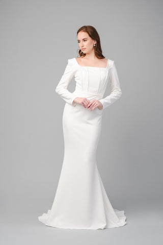 The Elea Dresses Louana dress in white is a fitted dress with long sleeves and a short train. The bodice is structured and features a square neckline.