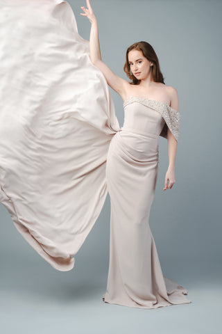 The Elea Dresses Quinn dress is strapless and features a beaded bust that extends over one shoulder. It is fitted, with a short train and an additional pleated train on the side.