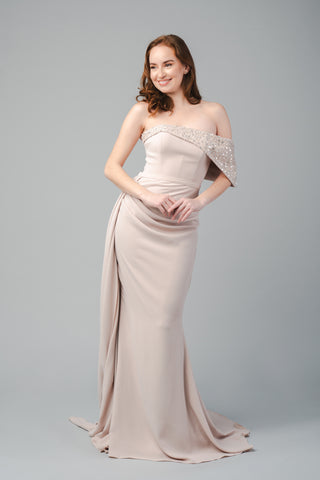 The Elea Dresses Quinn dress is strapless and features a beaded bust that extends over one shoulder. It is fitted, with a short train and an additional pleated train on the side.