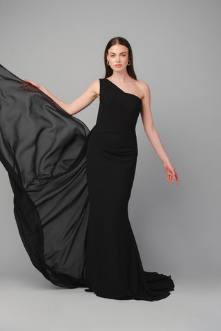 The Elea Dresses Selina dress in black is a one-shoulder fitted dress with a train. The top is pleated, and there is an additional pleated train on the side.