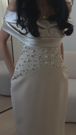 A model wears an elegant midi evening dress with a straight silhouette. Sparkling details adorn the center of the dress, catching the light with every subtle movement. The dress stays sleek and fitted, exuding sophistication from every angle.